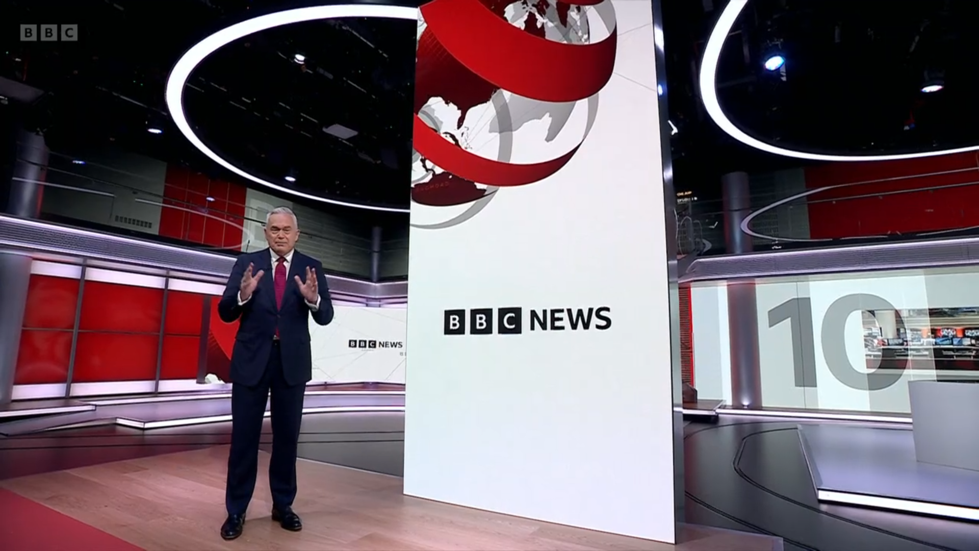 The new BBC News set in Studio B. It is a wide shot of a tall LED screen, with a large curved screen in the background, and part of the BBC logo as decoration up high above the set. Huw Edwards is standing in place. A large 10 is in the background